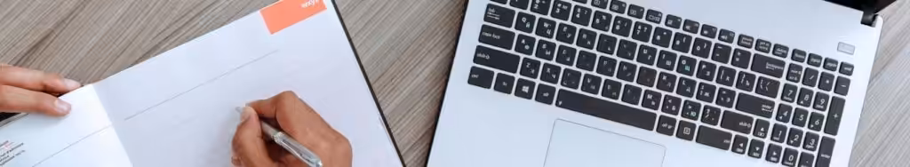 A person's hands engaged in work or study, with a laptop and a notebook visible on a wooden desk.